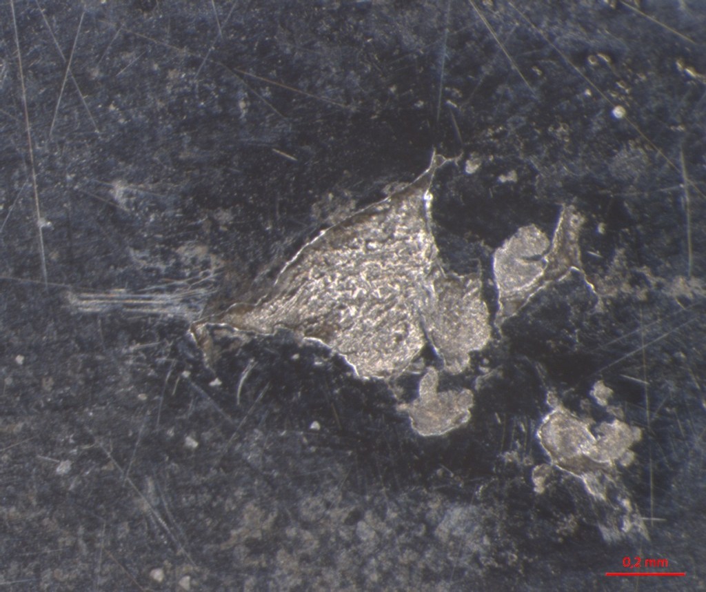 Microscopic view of metallic fragments or particles on a dark surface, with scale bar indicating 0.2 mm.